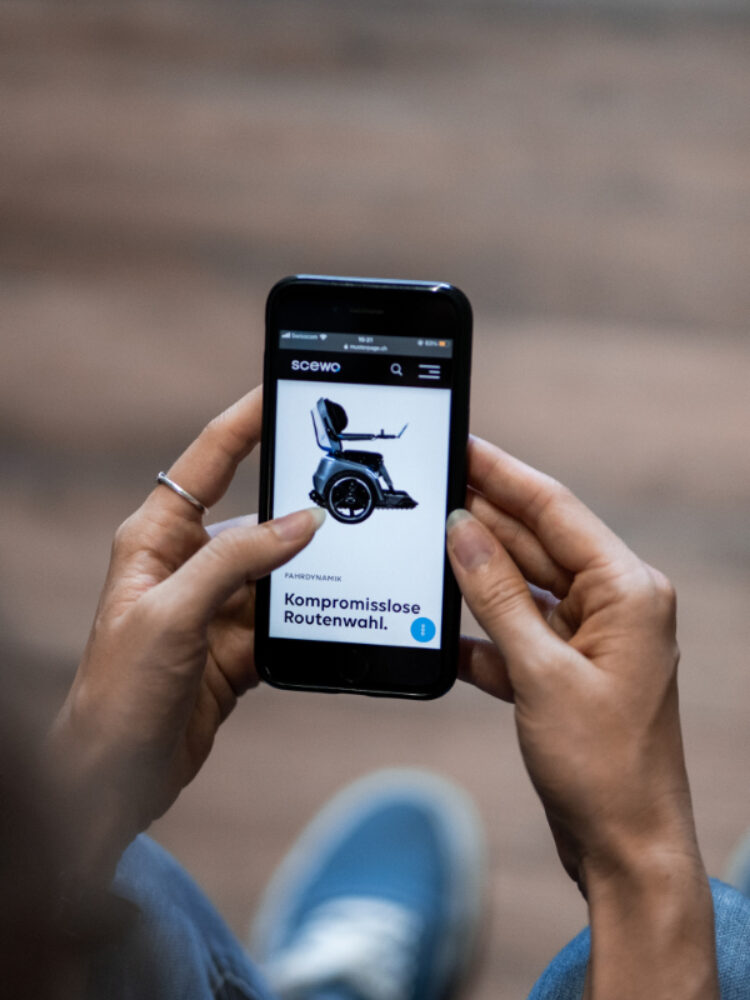 Hände halten ein Smartphone mit der Scewo Website und einer Abbildung des BRO Rollstuhls.