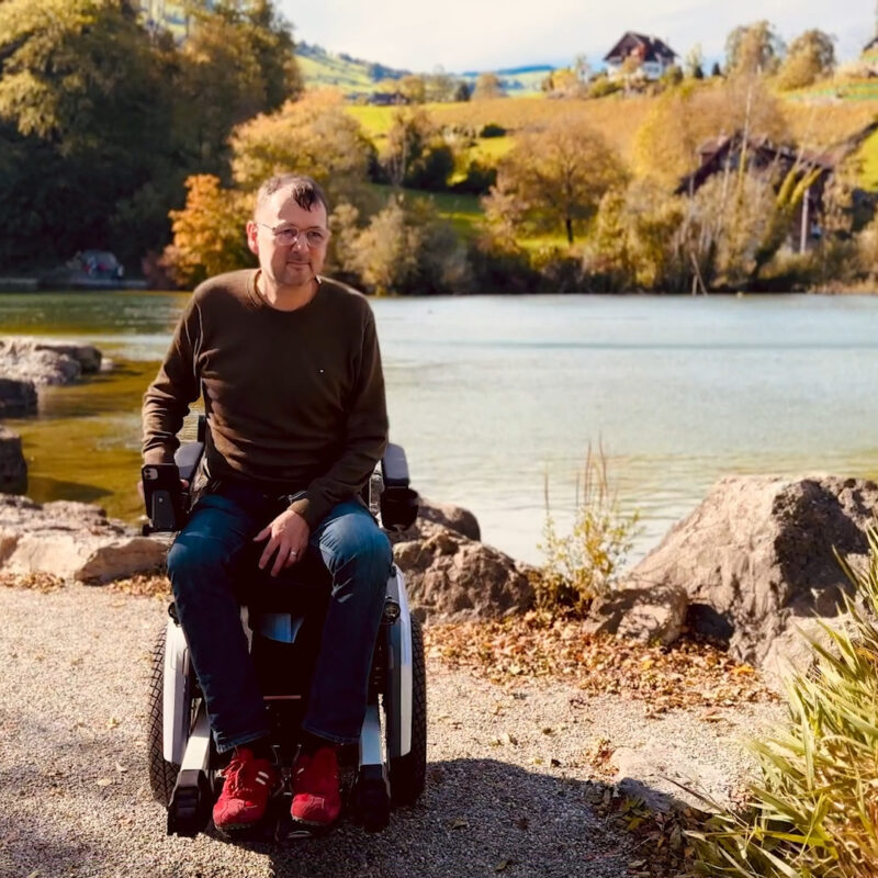 Michael sitzt in seinem Scewo BRO am Ufer eines Sees, umgeben von Felsen und herbstlicher Natur, während die Sonne auf das Wasser scheint. Im Hintergrund sind traditionelle Schweizer Häuser zu sehen.
