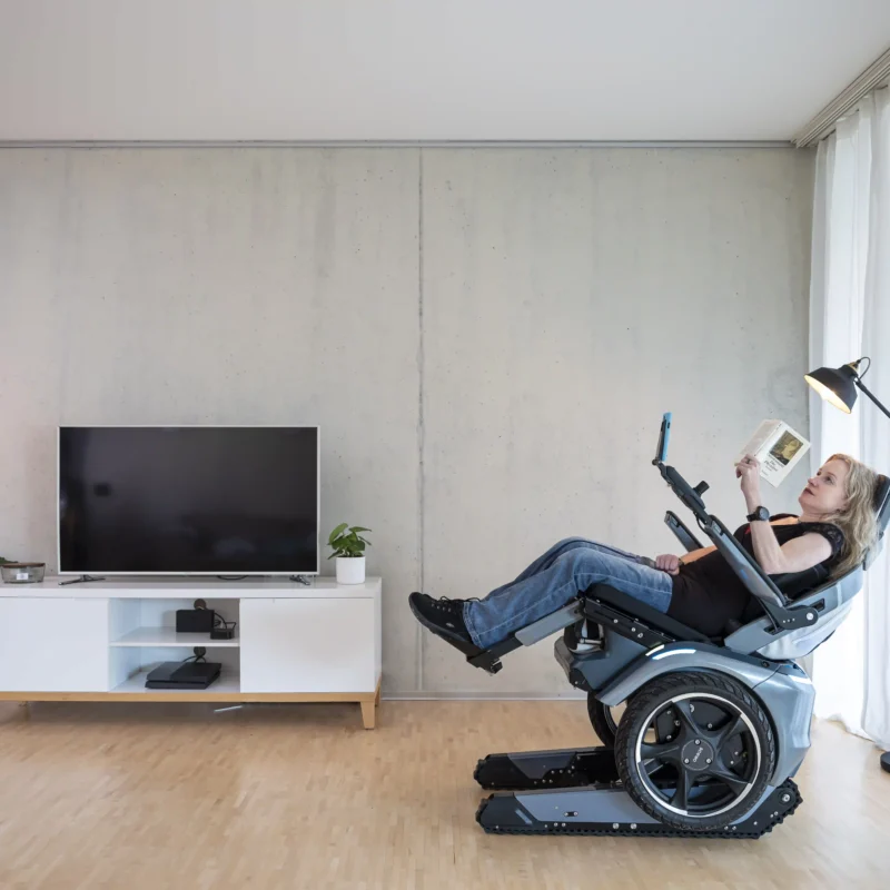 Eine Frau sitzt entspannt in der Relax-Position des Scewo BRO Elektrorollstuhls in einem modernen Wohnzimmer. Der Rollstuhl ist nach hinten geneigt, während sie ein Buch liest. Im Hintergrund stehen ein Fernseher, Pflanzen und eine Stehlampe, die eine gemütliche Atmosphäre schaffen.