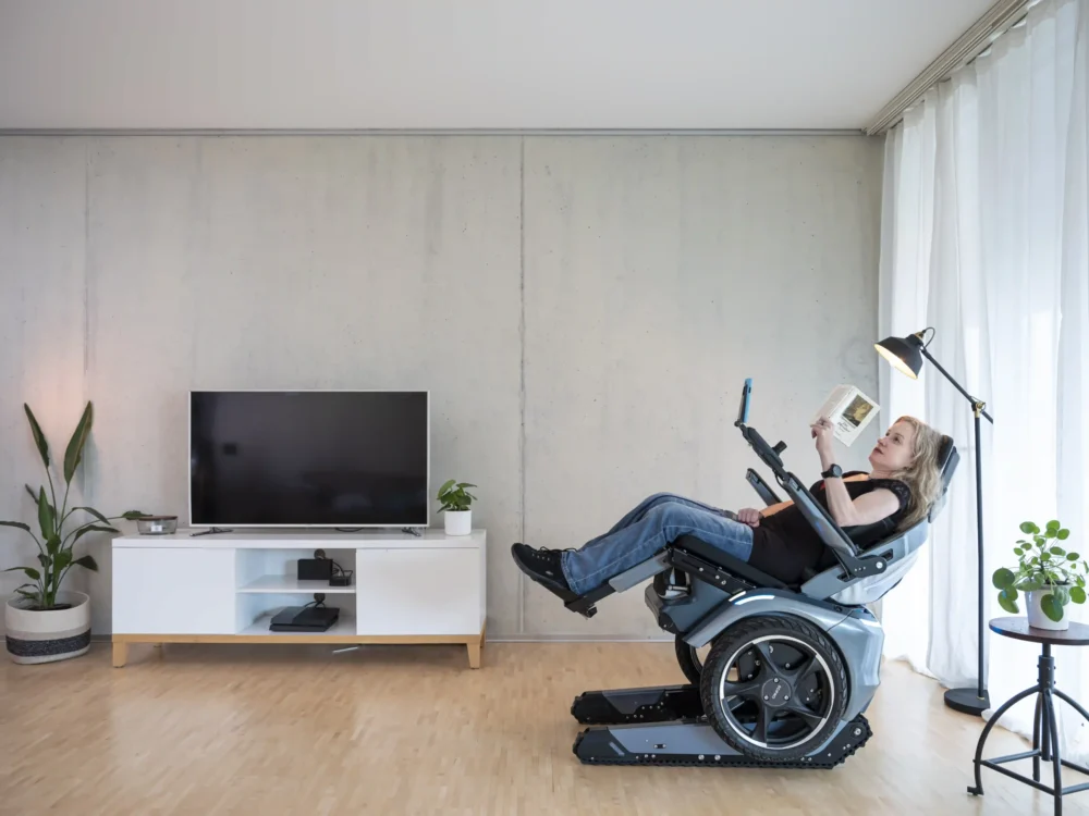 Eine Frau sitzt entspannt in der Relax-Position des Scewo BRO Elektrorollstuhls in einem modernen Wohnzimmer. Der Rollstuhl ist nach hinten geneigt, während sie ein Buch liest. Im Hintergrund stehen ein Fernseher, Pflanzen und eine Stehlampe, die eine gemütliche Atmosphäre schaffen.