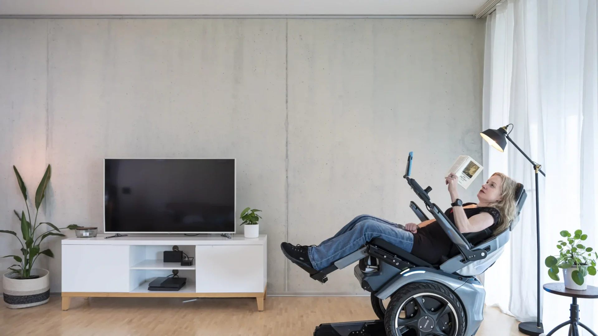 Eine Frau sitzt entspannt in der Relax-Position des Scewo BRO Elektrorollstuhls in einem modernen Wohnzimmer. Der Rollstuhl ist nach hinten geneigt, während sie ein Buch liest. Im Hintergrund stehen ein Fernseher, Pflanzen und eine Stehlampe, die eine gemütliche Atmosphäre schaffen.