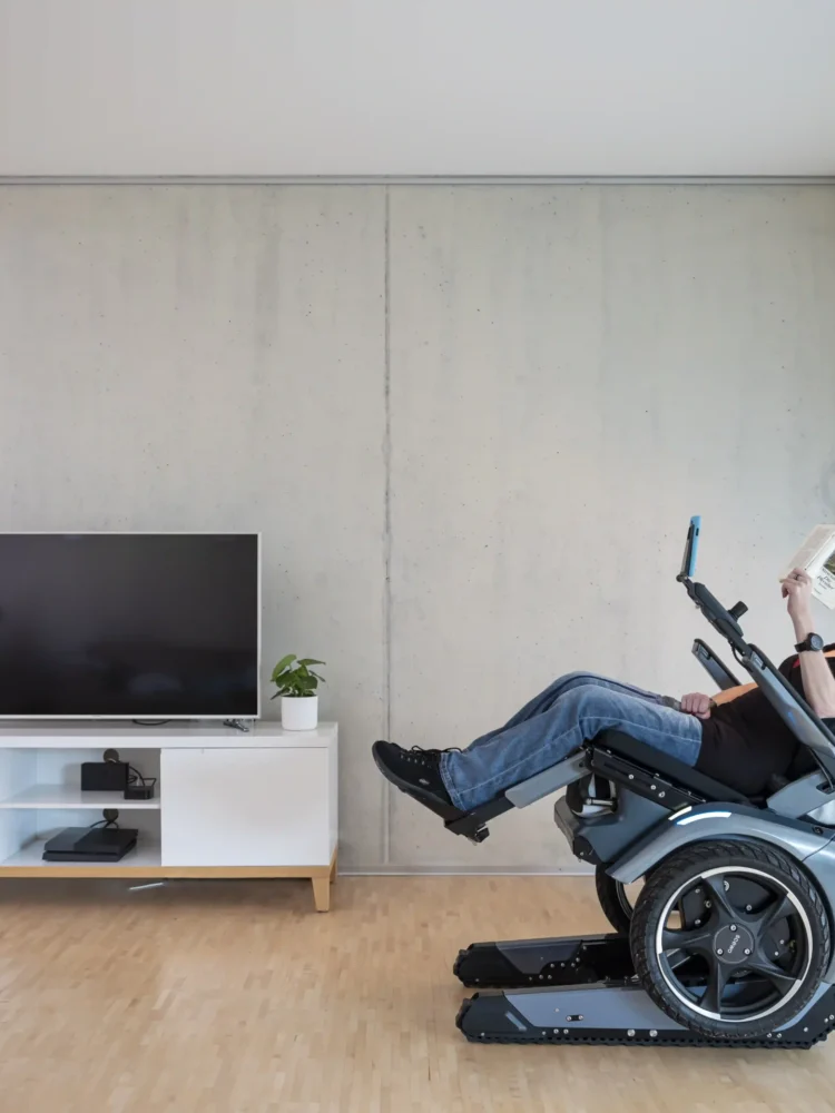 Eine Frau sitzt entspannt in der Relax-Position des Scewo BRO Elektrorollstuhls in einem modernen Wohnzimmer. Der Rollstuhl ist nach hinten geneigt, während sie ein Buch liest. Im Hintergrund stehen ein Fernseher, Pflanzen und eine Stehlampe, die eine gemütliche Atmosphäre schaffen.