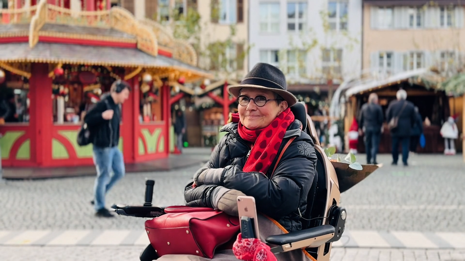 Anne sitzt in ihrem Scewo BRO vor einem Weihnachtsmarkt und lächelt. Sie trägt einen roten Schal, eine rote Tasche und einen Hut.