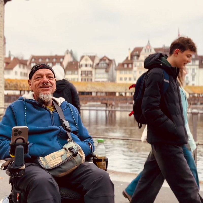 Stefan Baumgartner lächelt während eines Interviews mit dem Scewo BRO vor der Kappelbrücke in Luzern. Menchen laufen hinter ihm vrbei.