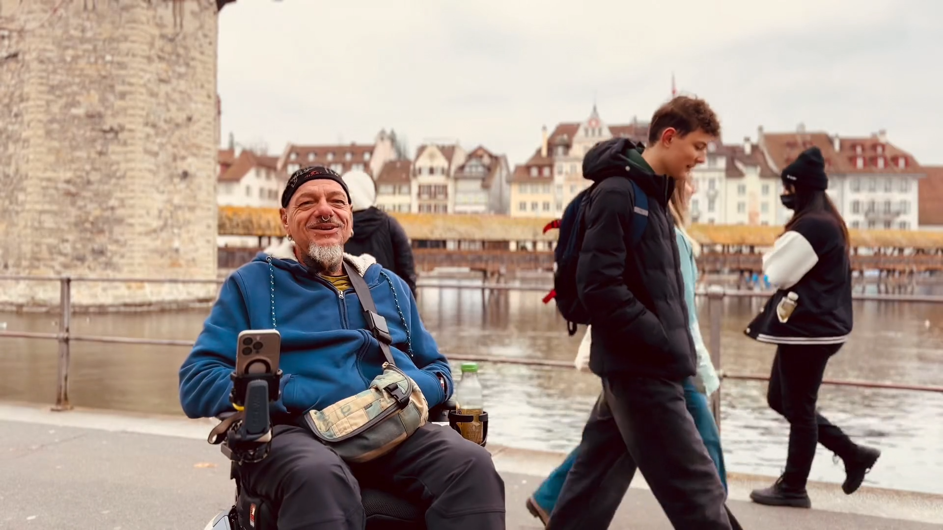 Stefan Baumgartner lächelt während eines Interviews mit dem Scewo BRO vor der Kappelbrücke in Luzern. Menchen laufen hinter ihm vrbei.