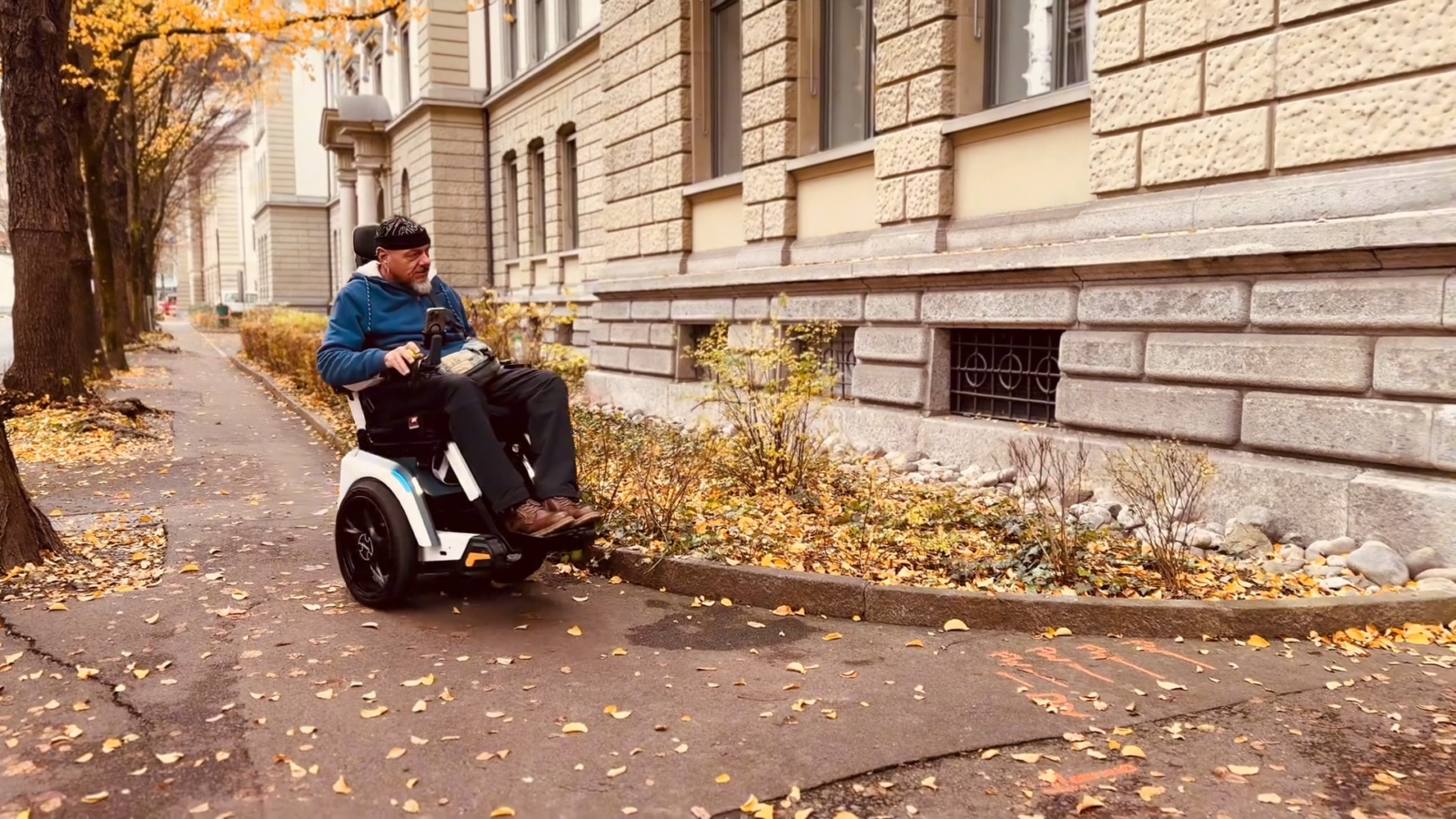 Stefan Baumgartner fährt mit dem ElektroollstuhlScewo BRO sicher und bequem auf einem Fußgängerweg mit Bäumen und einem großenGebäude im Hintergrund.