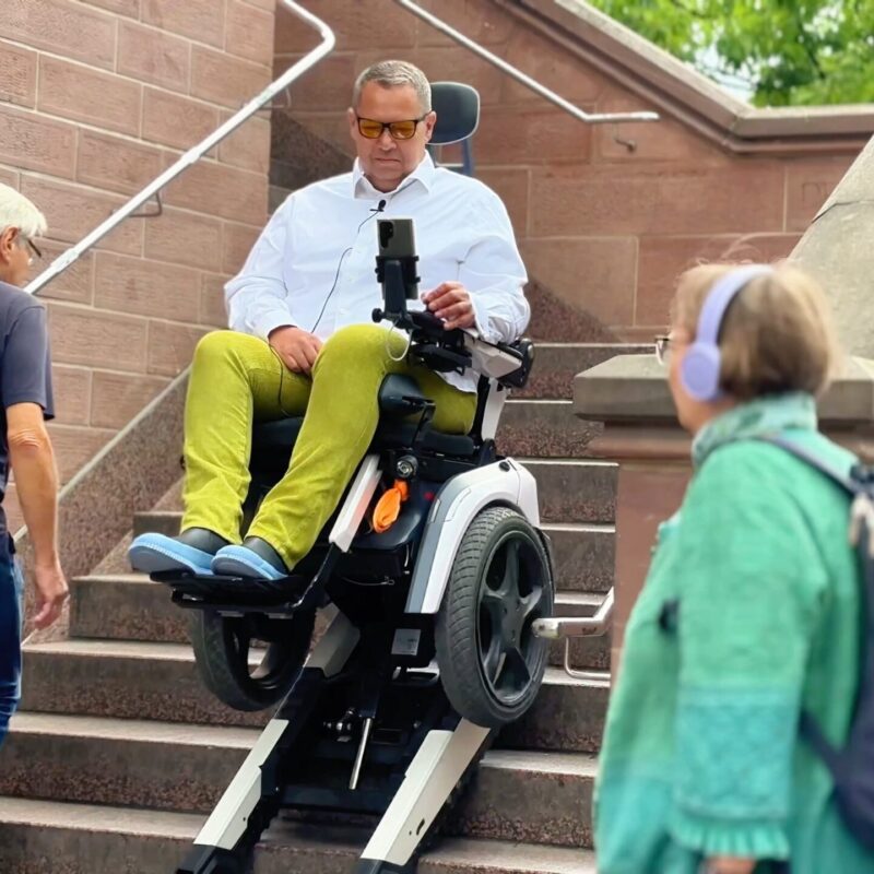 Daniel Fuks fährt in seinem treppensteigenden Elektrorollstuhl Scewo BRO auf dem eisernen Steg in Frankfurt die Treppen herunter