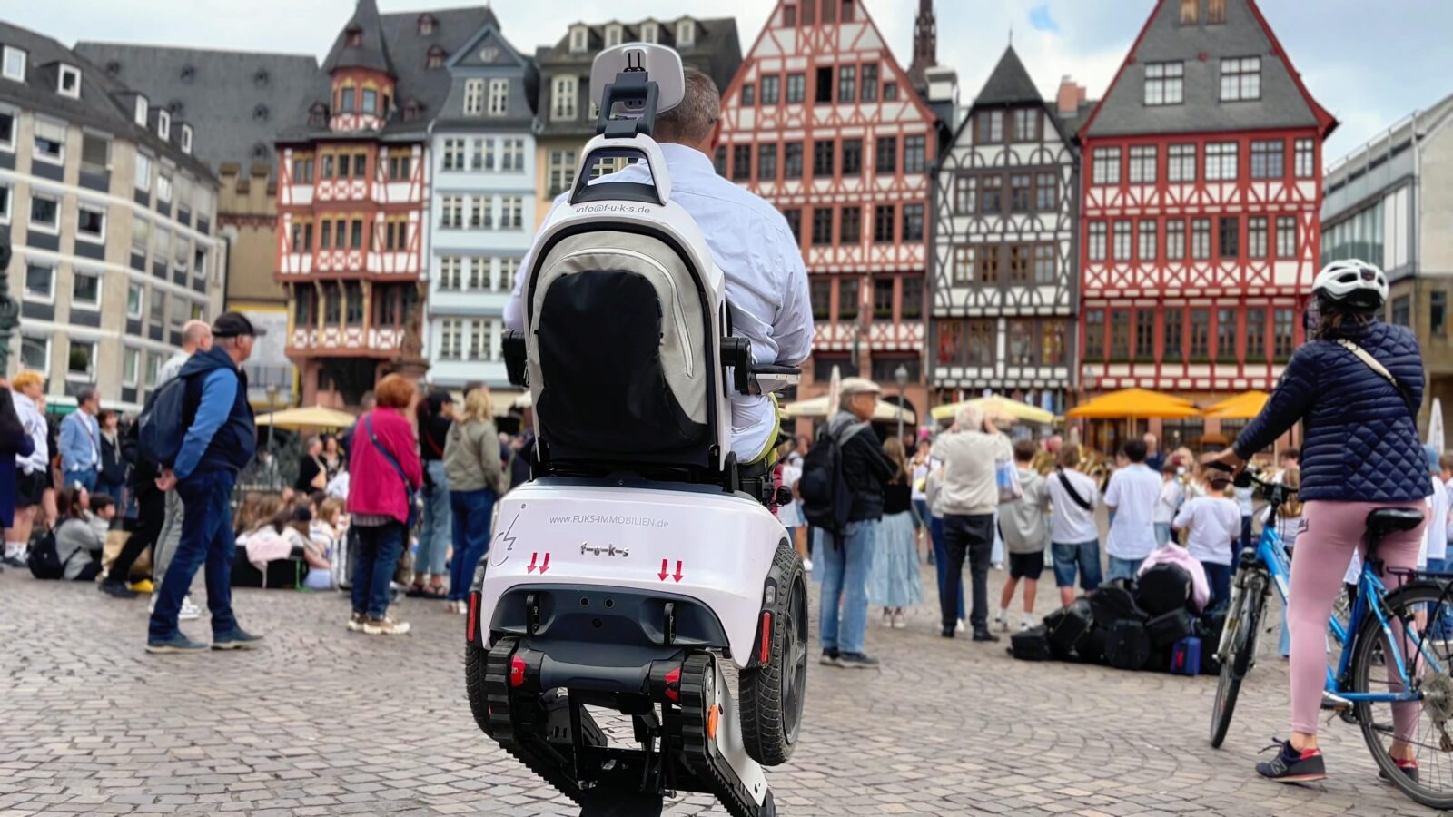 Daniel Fuks fährt den Sitz seines treppensteigenden Elektrorollstuhls Scewo BRO hoch vor der Altstadtkulisse Frankfurts mit einer großen Menschentraube im Hintergrund
