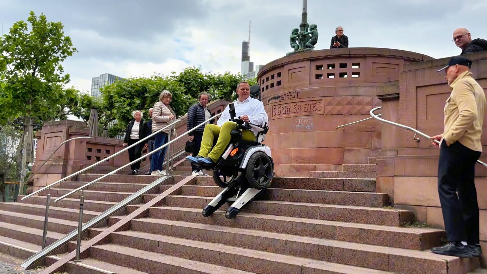 Daniel Fuks fährt in seinem treppensteigenden Elektrorollstuhl Scewo BRO auf dem eisernen Steg in Frankfurt die Treppen herunter und Menschen schauen ihm zu