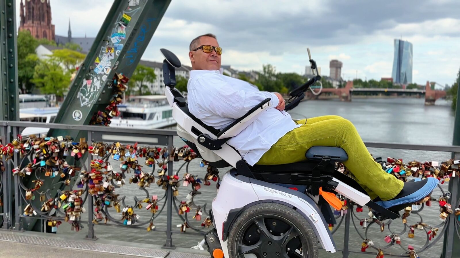 Daniel Fuks liegt in seinem treppensteigenden Elektrorollstuhl Scewo BRO im Relaxmodus beim eisernen Steg in Frankfurt und lächelt in die Kamera.
