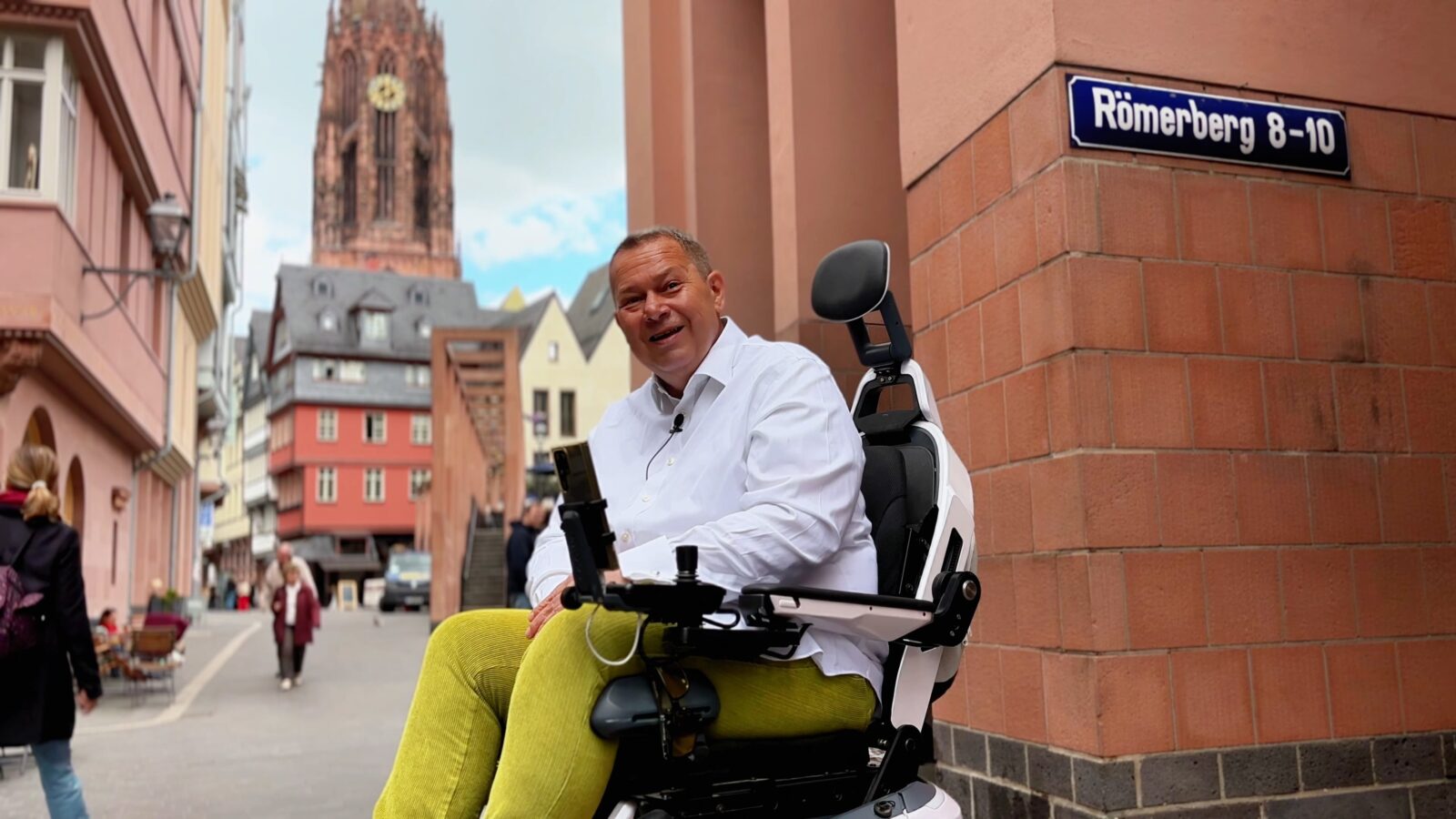 Daniel Fuks sitzt in seinem treppensteigenden Elktrorollstuhl Scewo BRO vor der Altstadtkulisse Frankfurts und lacht in die Kamera.