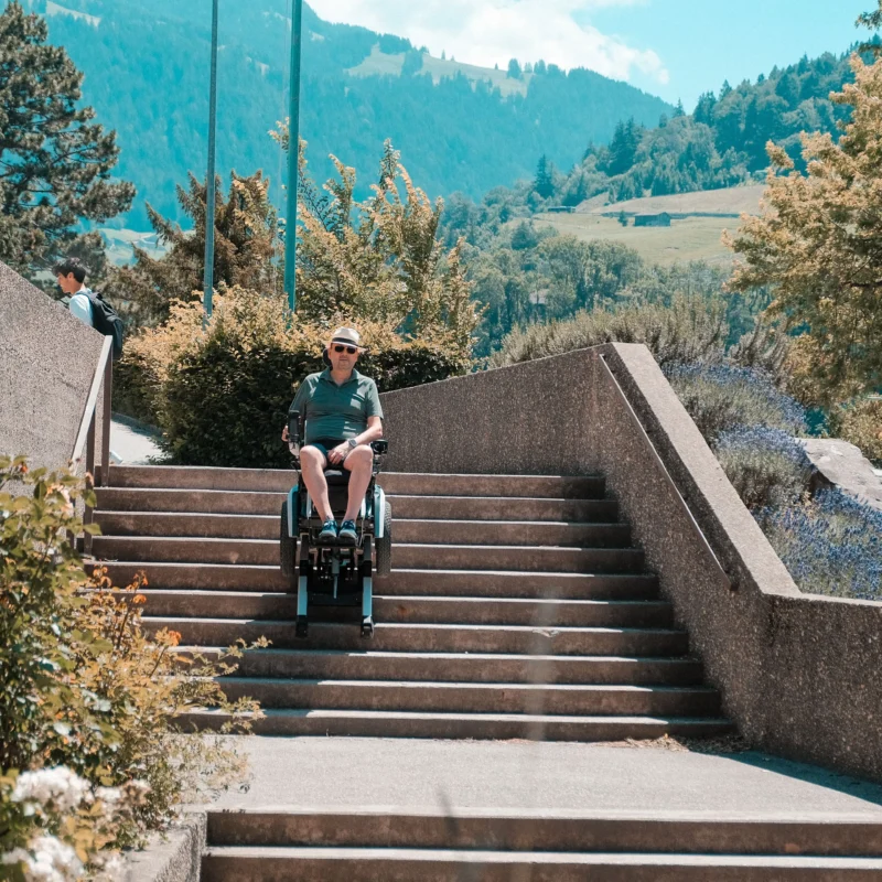 Mann fährt im Scewo BRO Elektrorollstuhl die Treppen hinunter. Im Hintergrund Berge und Bäume.
