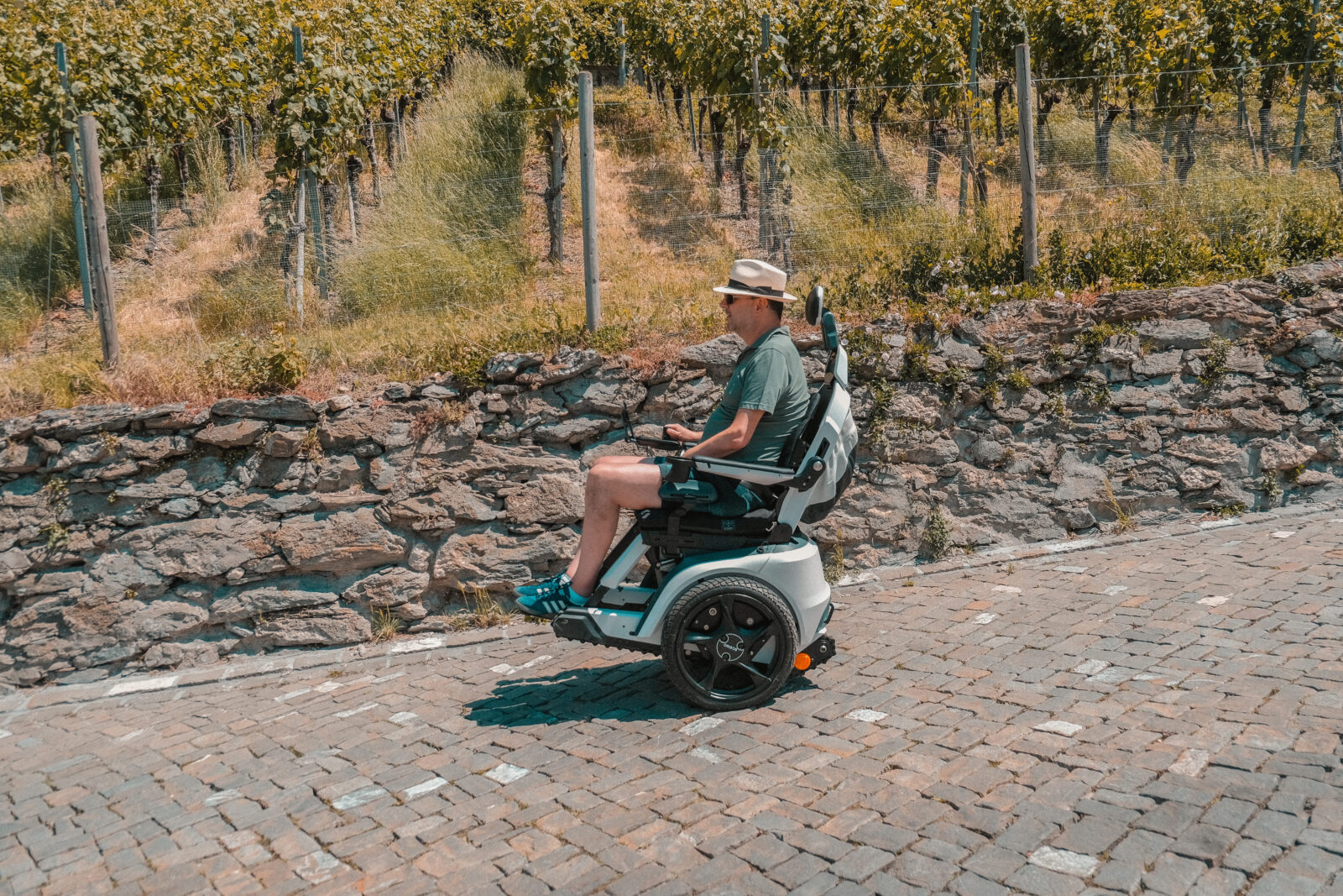 Ein Mann mit Hut fährt mit einem Scewo BRO auf einer gepflasterten Straße entlang einer Steinmauer, im Hintergrund Weinberge an einem sonnigen Tag.
