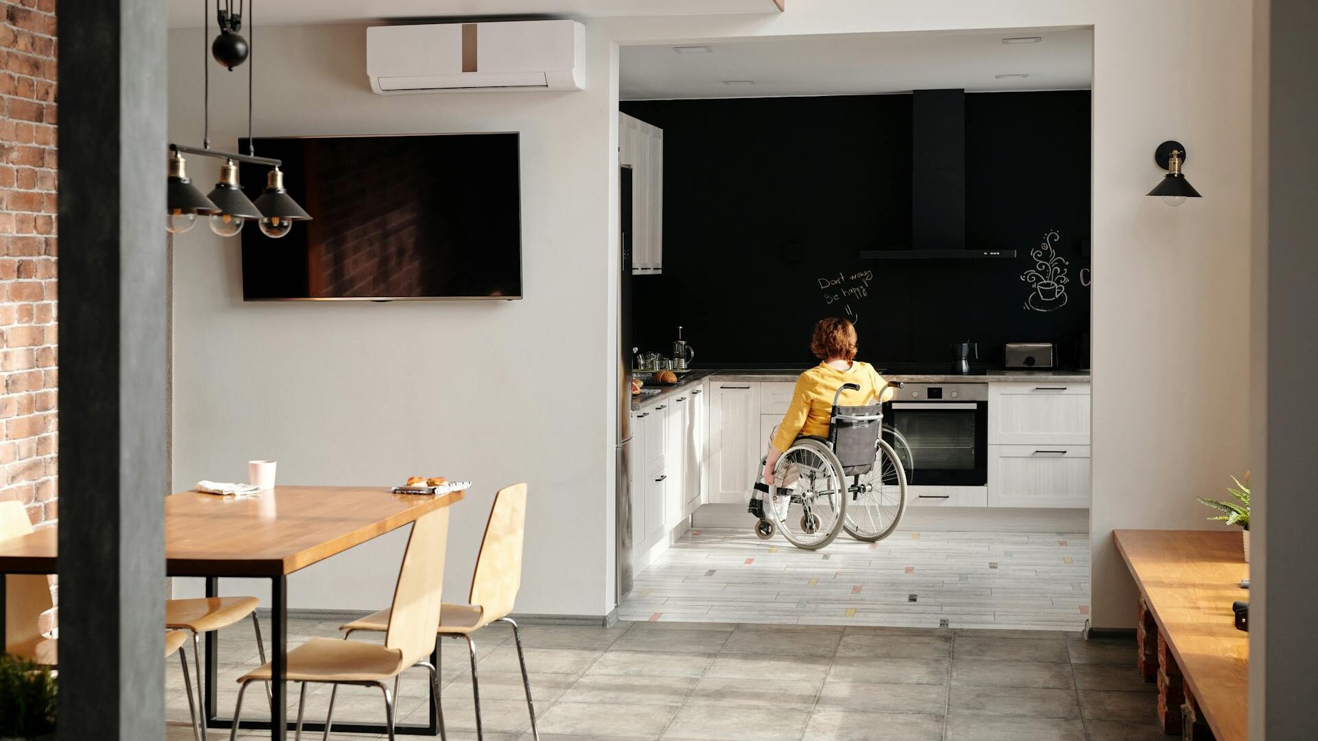 Person im Rollstuhl fährt durch eine moderne, helle Küche; im Vordergrund ein Esstisch mit Laptop, im Hintergrund schwarze Küchenwand mit Kaffeemaschine.