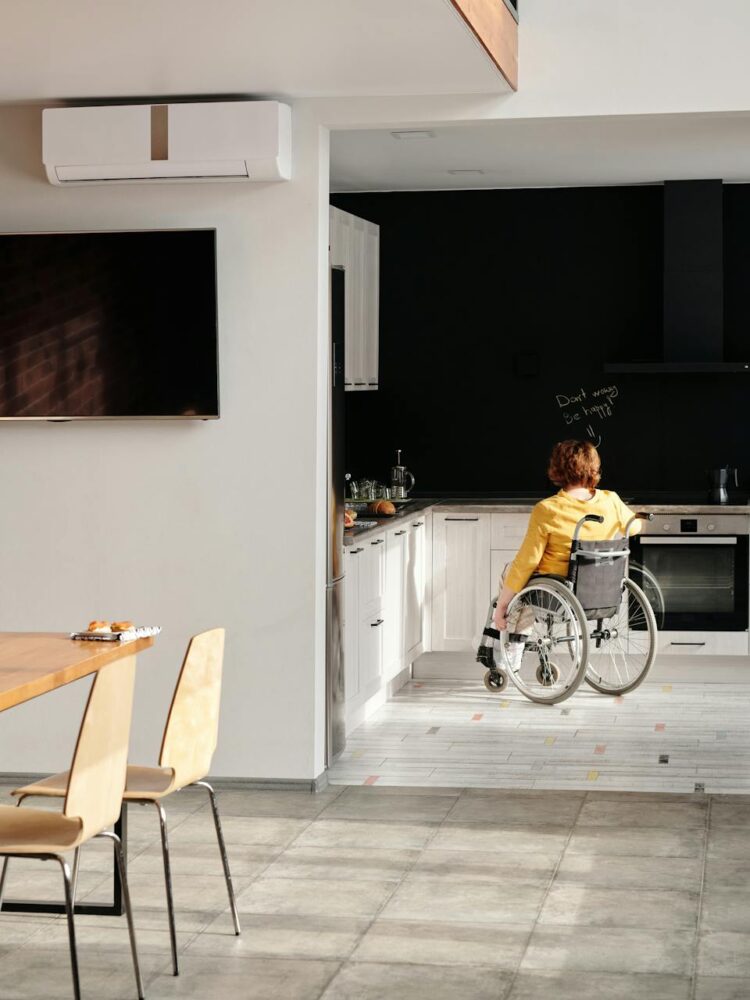 Person im Rollstuhl fährt durch eine moderne, helle Küche; im Vordergrund ein Esstisch mit Laptop, im Hintergrund schwarze Küchenwand mit Kaffeemaschine.