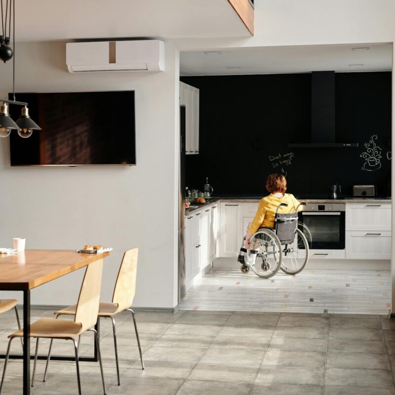 Person im Rollstuhl fährt durch eine moderne, helle Küche; im Vordergrund ein Esstisch mit Laptop, im Hintergrund schwarze Küchenwand mit Kaffeemaschine.