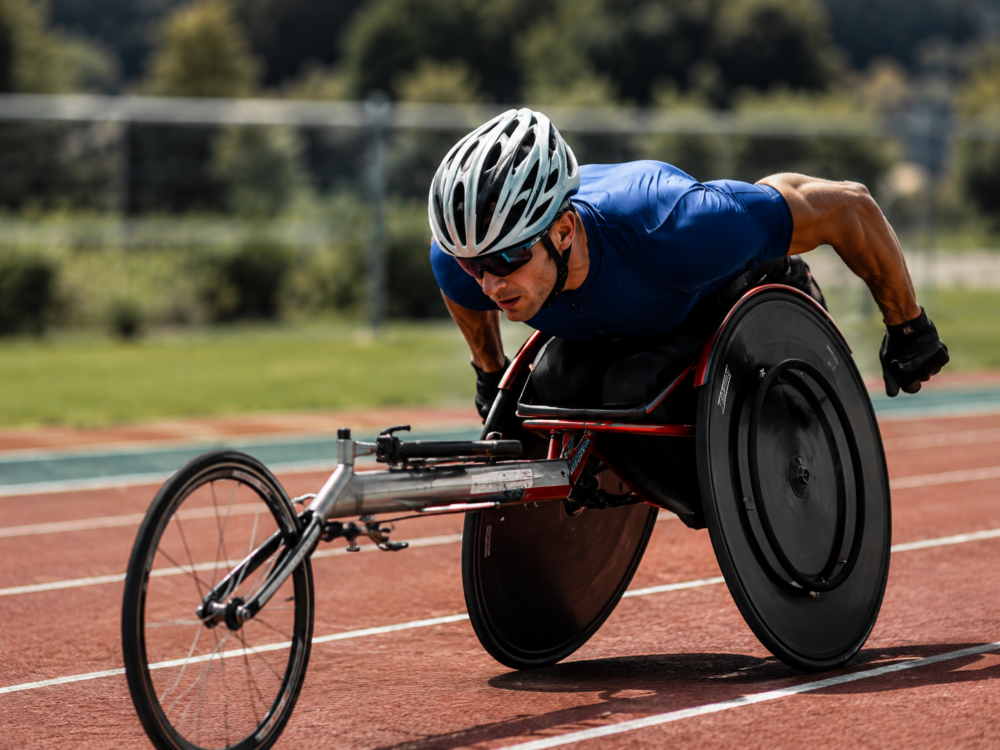 : Athlet im Rennrollstuhl fährt mit hoher Geschwindigkeit über die Laufbahn bei einem Para-Leichtathletik-Wettkampf.