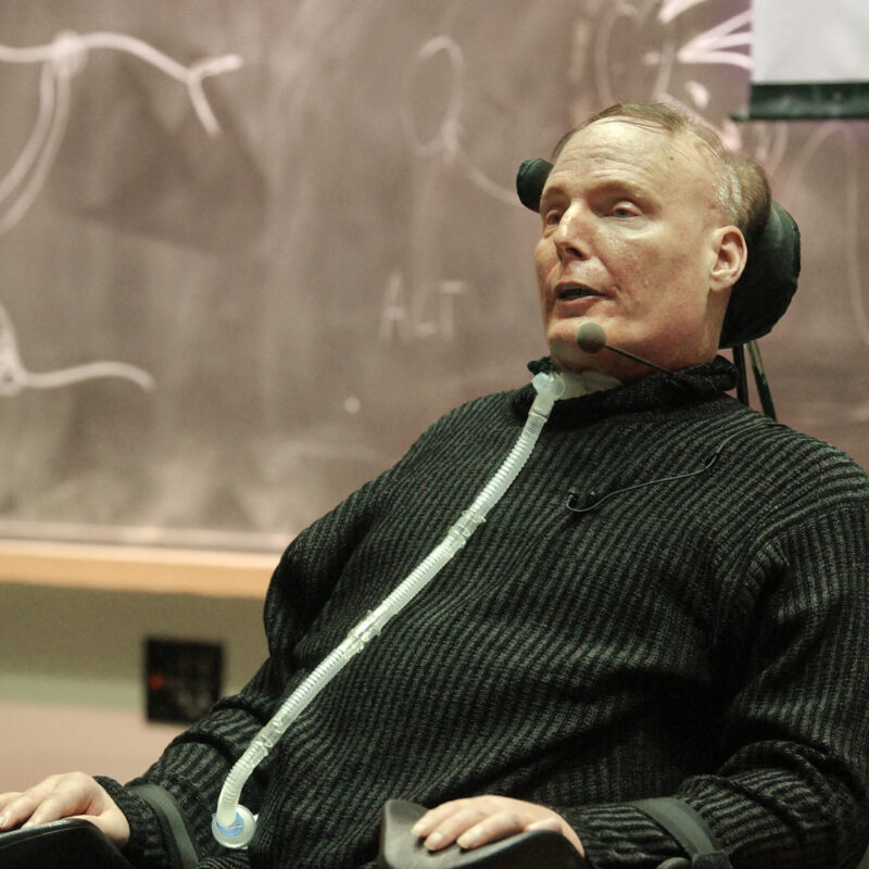 Christopher Reeve sitting in a wheelchair speaking into a microphone in front of a board