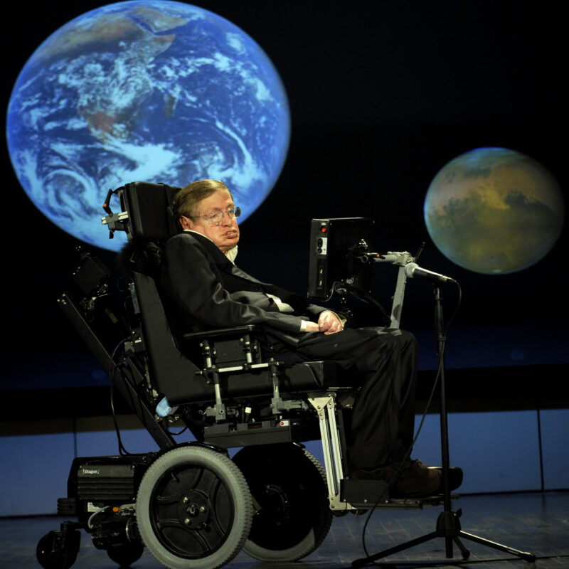 stephen hawking in his wheelchair in front of a big dark screen with two planets