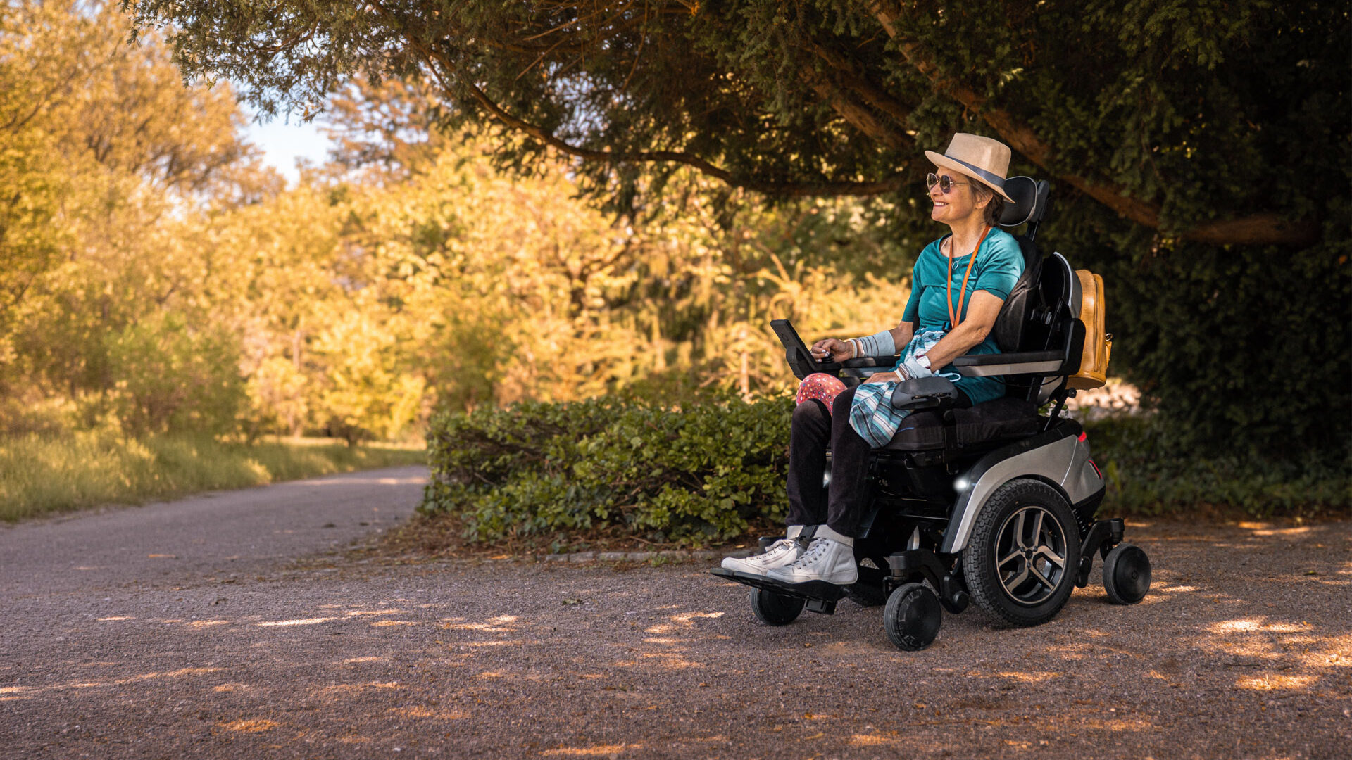 Lächelnde Frau mit Sonnenhut fährt im Scewo GO Elektrorollstuhl über einen Parkweg im Abendlicht.