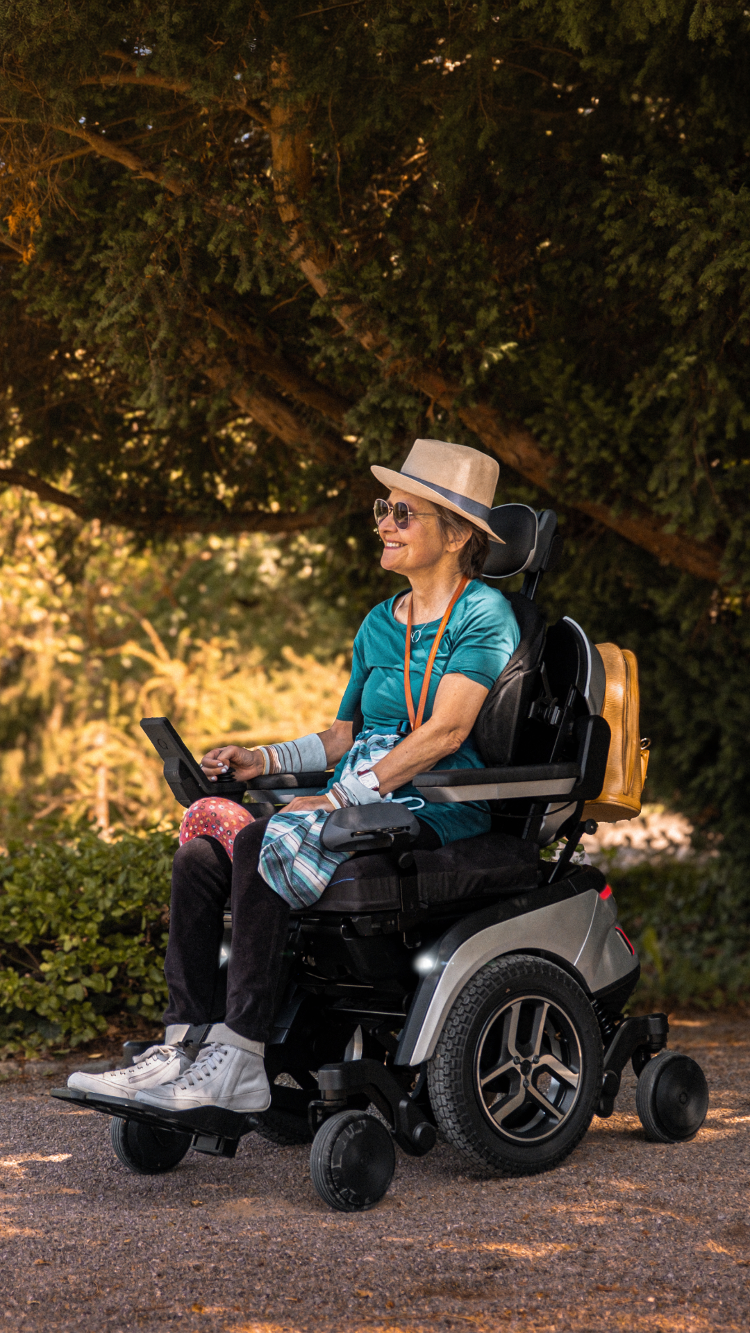 Lächelnde Frau mit Sonnenhut fährt im Scewo GO Elektrorollstuhl über einen Parkweg im Abendlicht.