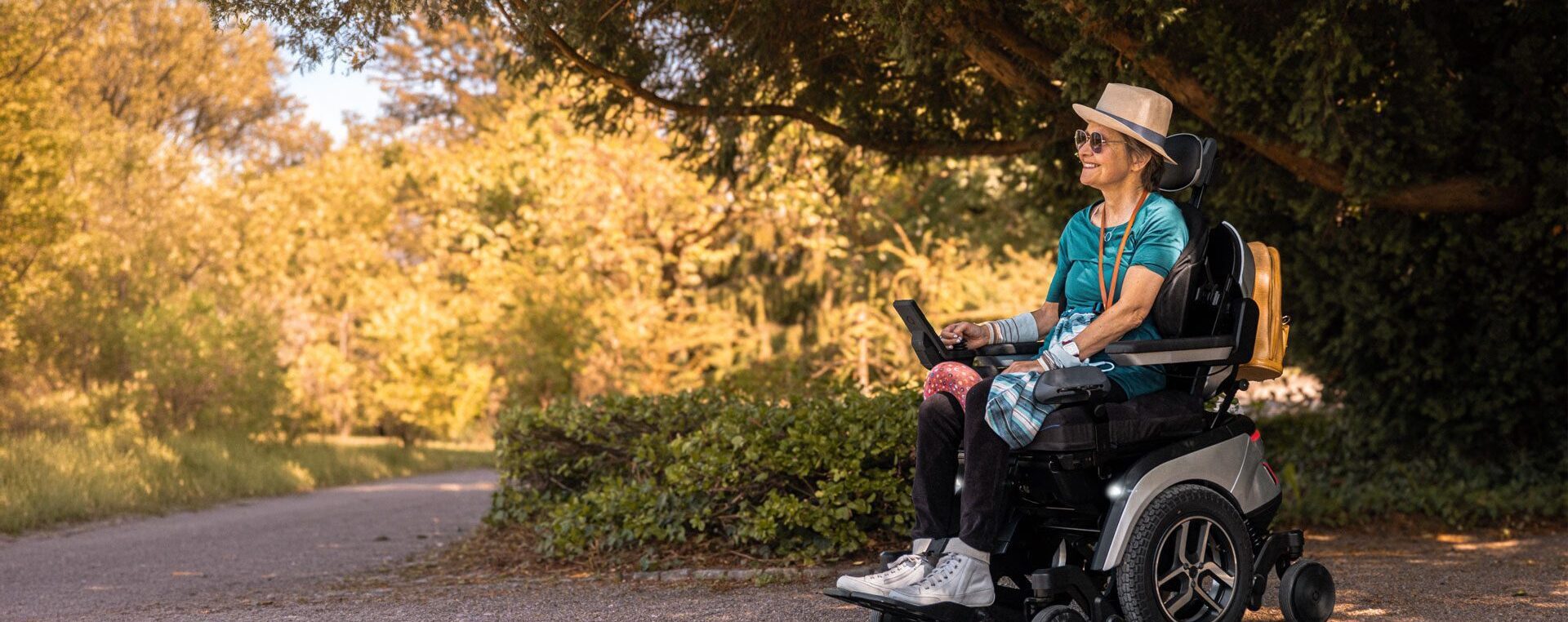 Lächelnde Frau mit Sonnenhut fährt im Scewo GO Elektrorollstuhl über einen Parkweg im Abendlicht.
