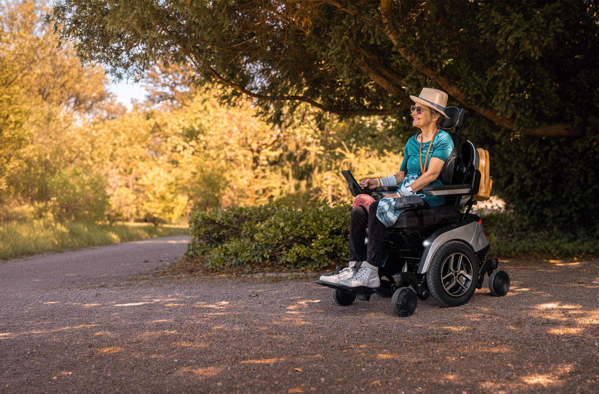 Lächelnde Frau mit Sonnenhut fährt im Scewo GO Elektrorollstuhl über einen Parkweg im Abendlicht.