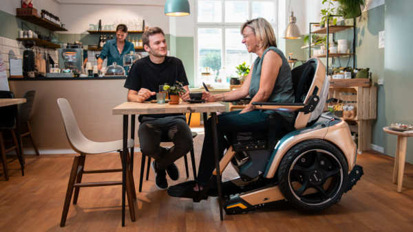 Frau im Scewo BRO mit abgesenktem Sitzlift beim Gespräch am Cafétisch.