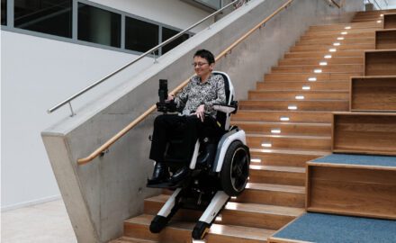 Frau fährt im Scewo BRO im Treppenmodus eine Holztreppe in einem Uni-Gebäude hinunter.