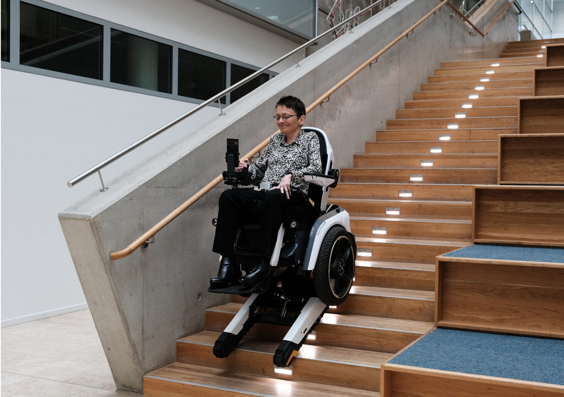Frau fährt im Scewo BRO im Treppenmodus eine Holztreppe in einem Uni-Gebäude hinunter.