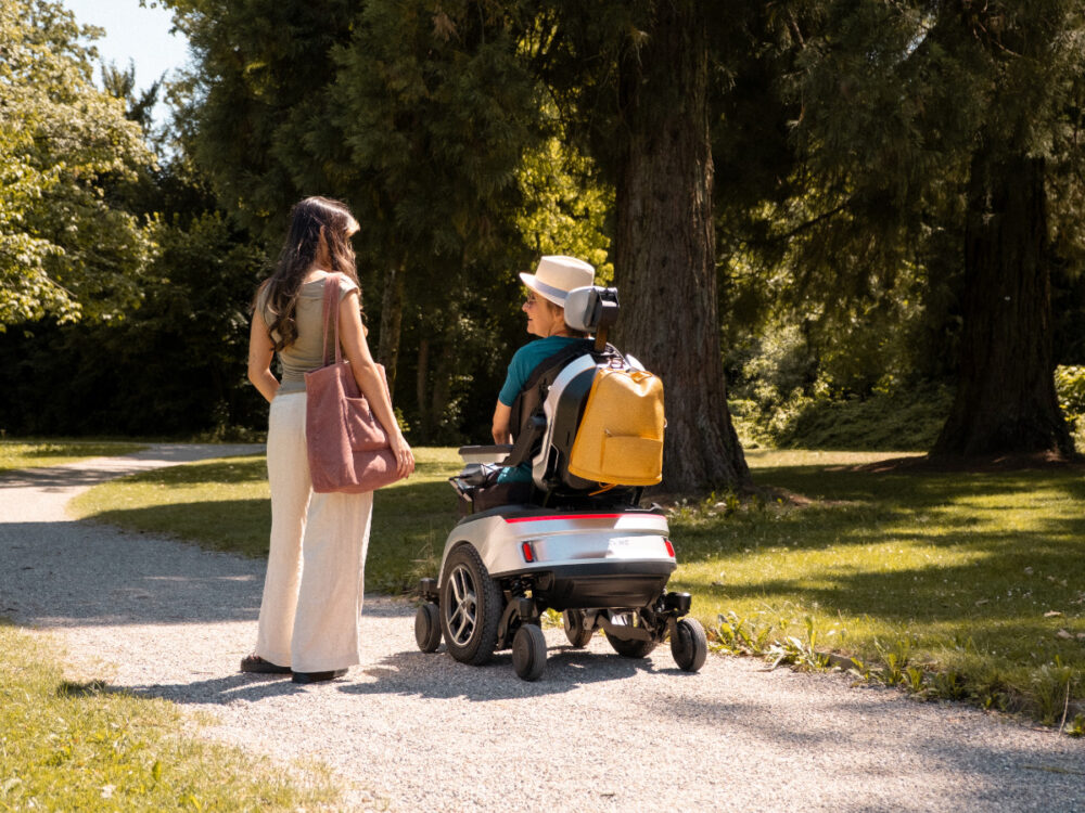 Zwei Frauen unterhalten sich auf einem Kiesweg im Park, eine sitzt im Scewo GO Elektrorollstuhl.
