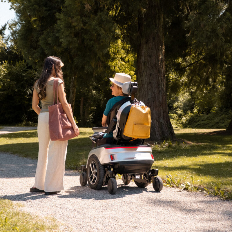 Zwei Frauen unterhalten sich auf einem Kiesweg im Park, eine sitzt im Scewo GO Elektrorollstuhl.